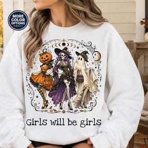 May include: A white long-sleeve sweatshirt featuring a graphic print of three figures: a pumpkin-headed character, a skeleton in a purple dress, and a ghost, all wearing witch hats. The text "Girls will be girls" is printed below the illustration.