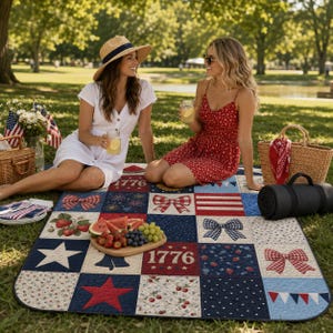 Puede incluir: Una manta de picnic de patchwork con diseños patrióticos, incluyendo estrellas, lazos y el número "1776". Dos personas están sentadas en la manta sobre la hierba. También hay una cesta de picnic, comida y bebidas.
