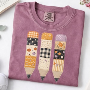 May include: A mauve short-sleeved t-shirt featuring a graphic of three pencils. Each pencil is composed of various patterned fabric segments including pumpkins, a ghost, flowers, gingham, and stripes. The shirt has a Comfort Colors brand label.