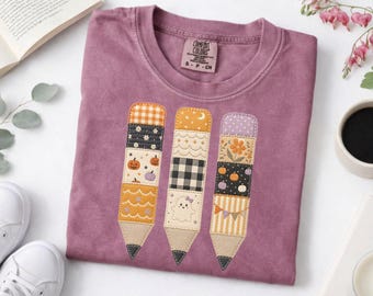 May include: A mauve short-sleeved t-shirt featuring a graphic of three pencils. Each pencil is composed of various patterned fabric segments including pumpkins, a ghost, flowers, gingham, and stripes. The shirt has a Comfort Colors brand label.