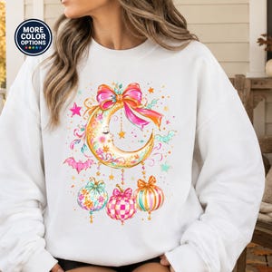 May include: A white long-sleeve sweatshirt featuring a graphic print of a crescent moon with a face, adorned with a large pink bow, hanging stars, small bats, and three colorful patterned pumpkins in shades of pink, blue, and orange.