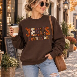 Peut inclure: Un sweat-shirt à col rond marron foncé avec l'inscription "JESUS loves you" dans une police décorative. Les lettres sont remplies de motifs sur le thème de l'automne, notamment des citrouilles orange, des feuilles, des fantômes et des carreaux.