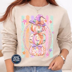 May include: A beige crewneck sweatshirt featuring a graphic of three stacked pumpkins. The top pumpkin wears a purple witch hat with a bow. The design includes pastel-colored vertical stripes, stars, and a circular border. A small circular badge reads MORE COLOR OPTIONS.