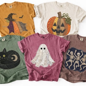 May include: Five short-sleeved t-shirts featuring Halloween-themed patchwork graphics. Designs include a witch on a mustard shirt, a pumpkin on cream, a black cat on sage green, a ghost on maroon, and dancing skeletons on a brown shirt.