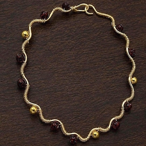 May include: A gold-colored necklace with a wavy design. The necklace features dark red beads and small gold-colored beads. The wire is wrapped around the beads and forms a unique, handcrafted piece of jewelry.