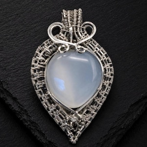 May include: A handcrafted silver pendant with a heart-shaped, pale blue gemstone. The gemstone is encased in a woven wire frame with decorative swirls at the top. The pendant is displayed on a dark, textured surface.