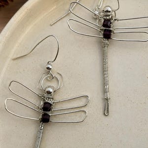 Può includere: Un paio di orecchini a forma di libellula realizzati in filo metallico argentato. Ogni orecchino presenta un lungo corpo in filo avvolto, quattro delicate ali modellate con il filo e due piccole perline quadrate viola scuro.