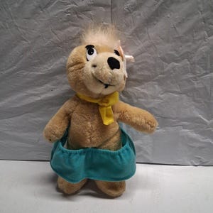 Pode incluir: Um brinquedo de pelúcia que lembra um urso castanho-claro com uma mecha de cabelo claro na cabeça, um pequeno laço rosa, um lenço amarelo no pescoço e uma saia azul-petróleo. O urso tem olhos pretos grandes, nariz preto e boca costurada.