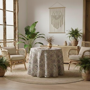 Puede incluir: Un salón acogedor con una mesa redonda cubierta con un mantel floral. Dos sillas de mimbre con cojines beige flanquean la mesa. Una gran planta en maceta y un tapiz de macramé completan la escena.