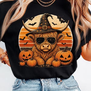 Puede incluir: Una camiseta negra con un gráfico de una vaca Highland con sombrero de bruja y gafas de sol. La vaca está centrada frente a un fondo de atardecer con murciélagos volando y tres calabazas de Halloween situadas en la base del diseño.