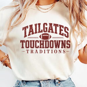 Puede incluir: Una camiseta de manga corta color crema con un estampado gráfico de color rojo oscuro con efecto desgastado. El texto dice TAILGATES, TOUCHDOWNS y TRADITIONS, con una ilustración de un balón de fútbol americano centrada entre las líneas.