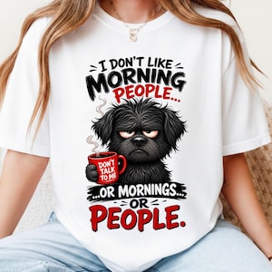 以下が含まれることがあります： DON'T TALK TO MEと書かれた赤いマグカップを持つ不機嫌そうな黒い犬のグラフィックがプリントされた白い半袖Tシャツ。周囲のテキストには、黒と赤の文字でI DON'T LIKE MORNING PEOPLE... OR MORNINGS... OR PEOPLEと書かれています。