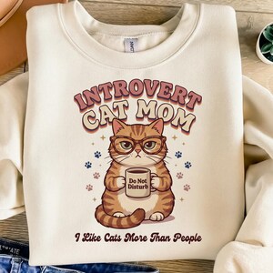 Puede incluir: Sudadera color crema con el texto "Introvert Cat Mom" y un gato de dibujos animados con gafas que sostiene una taza con "Do Not Disturb". La frase "I Like Cats More Than People" está debajo.