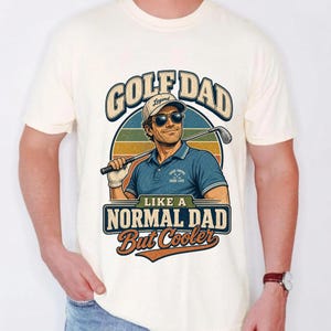 May include: A cream-colored t-shirt featuring a graphic of a person wearing a cap and sunglasses, holding a golf club. The text reads GOLF DAD LIKE A NORMAL DAD But Cooler. The design includes a circular background with horizontal stripes in muted colors.