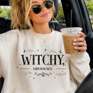 May include: A cream-colored crewneck sweatshirt featuring black text that reads WITCHY, OBVIOUSLY. The design includes decorative black stars, crescent moons, and delicate brown leaf sprigs surrounding the lettering on the front of the garment.