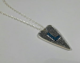 May include: A silver necklace with a silver heart pendant. The heart is covered in silver glitter and has a blue glitter letter "L" in the center.