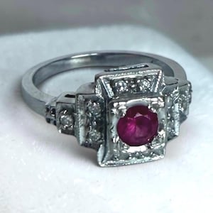 May include: A silver ring with a square setting featuring a vibrant, round, ruby gemstone. The setting is embellished with small, clear accent stones. The ring has a classic, vintage design.