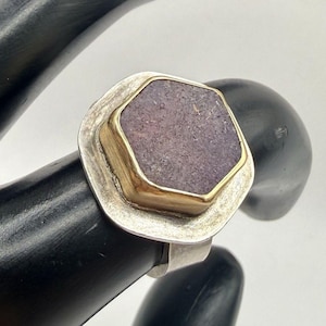 Könnte beinhalten: Ein Ring mit einem hexagonalen, texturierten violetten Stein in einer goldfarbenen Fassung. Der Stein ist auf einer breiten, flachen, silberfarbenen Metallbasis mit gebürsteter Oberfläche montiert. Die Ringschiene ist glatt und silberfarben.