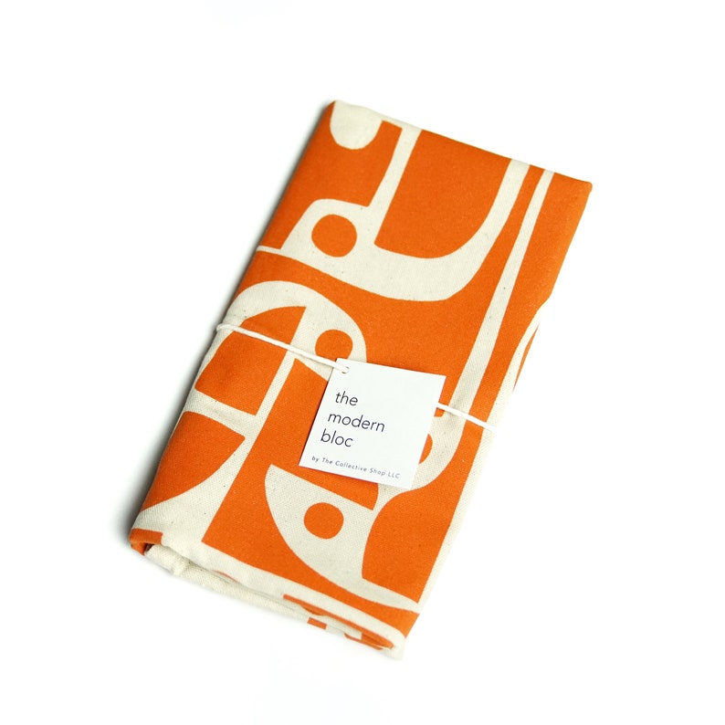 Op de afbeelding: Oranje en witte keukendoek met een geometrisch patroon. De doek is gevouwen en vastgebonden met een touwtje. Een wit label is bevestigd aan de doek met de tekst "the modern bloc by The Collective Shop LLC".