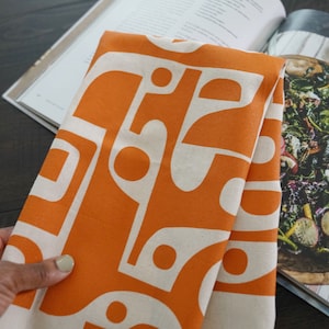 Op de afbeelding: Oranje keukendoek met een wit geometrisch patroon. De handdoek is gevouwen en ligt op een kookboek.