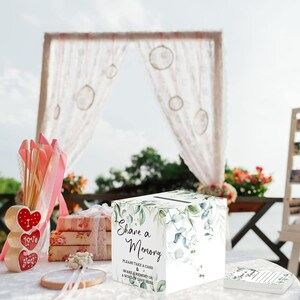 Puede incluir: Una caja de tarjetas blanca con una ranura en la parte superior, decorada con hojas verdes y el texto "Share a Memory". Otros artículos incluyen una decoración en forma de corazón de madera, una pila de regalos y un pequeño letrero con texto.