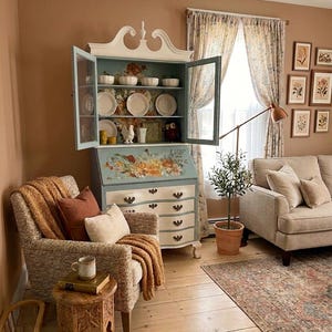 May include: A cozy living room scene featuring a blue and white cabinet with open glass doors displaying dishes. A patterned armchair with a knitted throw and pillows sits near a small wooden table. The room has a warm color palette with floral curtains and framed botanical prints.