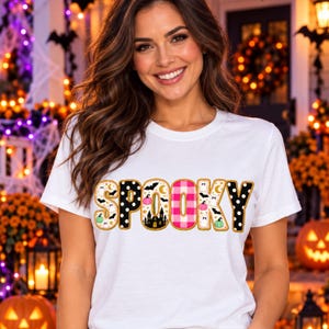 May include: A white short-sleeved t-shirt featuring the word SPOOKY printed across the chest. Each letter has a unique pattern including black and white polka dots, pink and white buffalo plaid, and a cream background with ghosts and pumpkins.