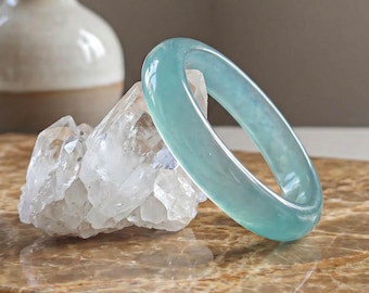 May include: A translucent, light blue jade bangle bracelet rests on a clear quartz crystal. The bracelet is smooth and circular, with a polished finish. The quartz sits on a brown marble surface, with a vase of dried flowers in the background.
