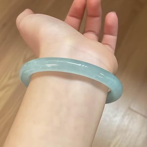 May include: A pale blue-green jade bangle bracelet. The smooth, rounded bracelet is translucent. The bangle is worn on a wrist.
