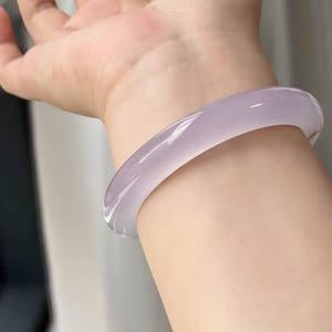 May include: A translucent, lavender-colored bangle bracelet. The smooth, polished surface reflects light, showcasing the subtle variations in color. This elegant piece of jewelry is a classic accessory.