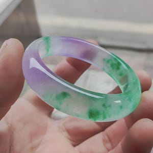 Puede incluir: Una pulsera de jade translúcida. La pulsera presenta un degradado de colores, incluyendo morado, verde y blanco. Las áreas verdes tienen un patrón moteado. La pulsera se sostiene en una mano, mostrando su superficie lisa y pulida.