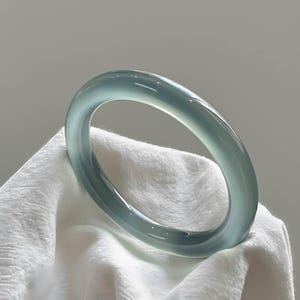 May include: A pale blue-green jade bangle bracelet. The smooth, polished surface reflects light, showcasing the natural variations in the stone. The bracelet is displayed on a white, textured fabric, highlighting its elegant design.