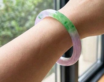 May include: A translucent jade bangle bracelet with a gradient of colors, including green and lavender. The bracelet is smooth and polished, showcasing the natural variations in the stone. The bracelet is worn on a wrist.