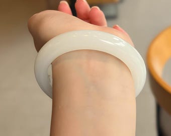 May include: A smooth, white jade bangle bracelet. The bracelet is a solid, circular band, showcasing a polished, glossy finish. The jewelry is worn on a wrist, with the skin tone contrasting against the bright white of the bracelet.