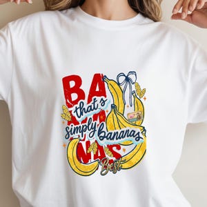 Könnte beinhalten: Weißes T-Shirt mit einem farbenfrohen Grafikdesign. Der Aufdruck zeigt den Text "BA that's simply Bananas MAS Best" in Rot, Blau und Gelb, ergänzt durch Bananen-Illustrationen und dekorative Elemente.