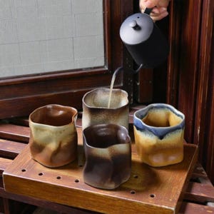 May include: Four ceramic cups with irregular shapes and earthy tones of brown, beige, and blue are arranged on a wooden tray. A black kettle pours liquid into one of the cups, suggesting a tea or coffee preparation.