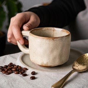 Puede incluir: Una taza de cerámica de color crema, de forma redondeada y con un asa pequeña. La taza está sobre un platillo a juego, junto a granos de café y una cuchara dorada. La taza tiene un aspecto rústico y texturizado.