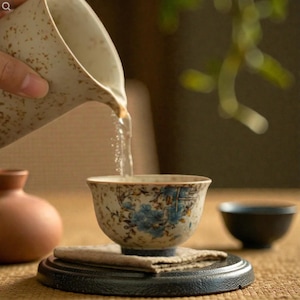 Puede incluir: Una taza de té de cerámica con diseños florales azules se está llenando de agua de una jarra a juego. La taza se encuentra sobre un pequeño posavasos sobre una base redonda negra. Un cuenco pequeño y oscuro está en el fondo.