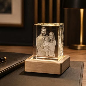 Op de afbeelding: Een rechthoekig glazen blok met een lasergegraveerd driedimensionaal portret van een gezin van drie personen, bestaande uit twee volwassenen en een kind. Het glazen blok is gemonteerd op een lichtgekleurde houten voet die het kristal verlicht.