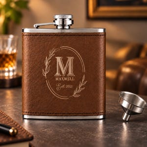 May include: A brown leather-wrapped stainless steel flask with a silver screw-top lid. The front features an engraved oval frame with a large letter M, the name MAXWELL, and the text Est. 2021, accompanied by a small silver metal funnel.