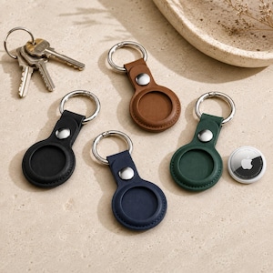 Peut inclure: Quatre porte-clés circulaires en cuir noir, marron, bleu marine et vert foncé, chacun doté d'un bouton-pression en métal argenté et d'un anneau. Un trousseau de clés en métal argenté et un traceur blanc avec le logo Apple sont présents.