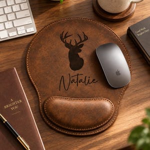 Peut inclure: Un tapis de souris en cuir marron avec repose-poignet, présentant une silhouette de tête de cerf gravée en marron foncé et le prénom Natalie en écriture cursive. Le tapis de souris a une forme arrondie avec des bords cousus et une surface lisse.