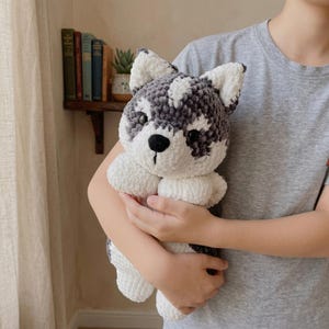 May include: A handmade plush husky dog with a black nose, black and grey markings, and white accents. The stuffed animal is made of soft, textured yarn and is being held.