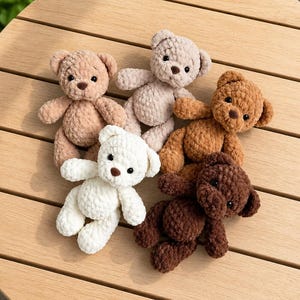 May include: A collection of five handmade crochet teddy bears in various colors: tan, light brown, beige, brown, and white. Each bear has small black eyes and a brown nose. The bears are arranged on a wooden table.