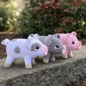May include: Three crocheted pig figurines in a row. The first pig is white with gray spots, the second is gray, and the third is pink. All have pink snouts, black eyes, and beige hooves. The pigs are standing on a stone surface.