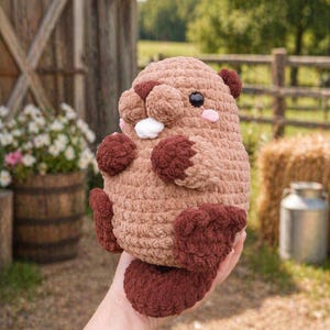 Puede incluir: Un juguete de castor de peluche hecho a mano, en tonos marrón y marrón oscuro. El animal de ganchillo tiene un cuerpo redondo, orejas pequeñas y un mechón de pelo blanco. El castor se sostiene en una mano, con un fondo borroso.