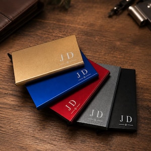 Puede incluir: Un conjunto de cinco tarjeteros metálicos rectangulares en oro, azul, rojo, gris y negro. Cada estuche presenta las letras grabadas JD sobre una pequeña forma de diamante y líneas horizontales. Los tarjeteros están dispuestos en abanico.