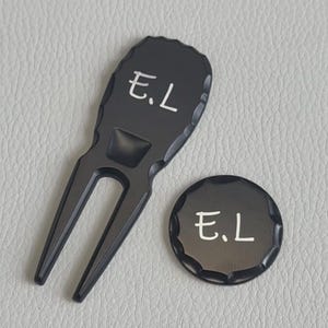 Könnte beinhalten: Ein mattes, schwarzes Golf-Pitchgabel-Werkzeug aus Metall und ein passender runder Ballmarker. Beide Gegenstände haben ein gewelltes Randdesign und sind mit der weißen Aufschrift E.L. graviert. Das Werkzeug hat eine zweizinkige Basis.
