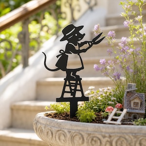 May include: A black metal garden stake silhouette featuring a mouse wearing a hat and apron, standing on a small ladder while holding a pair of garden shears. The mouse figure is positioned upright with a long tail and is designed for outdoor use.