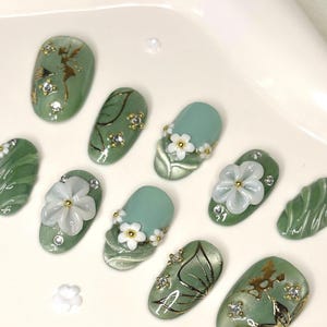 May include: A collection of ten artificial nails in shades of green and blue. Some nails are decorated with white flower accents, others with gold leaf and rhinestones. The nails are displayed on a white surface.